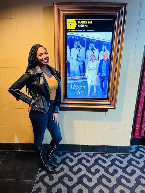 Smiling Kara standing next to Marry Me movie poster